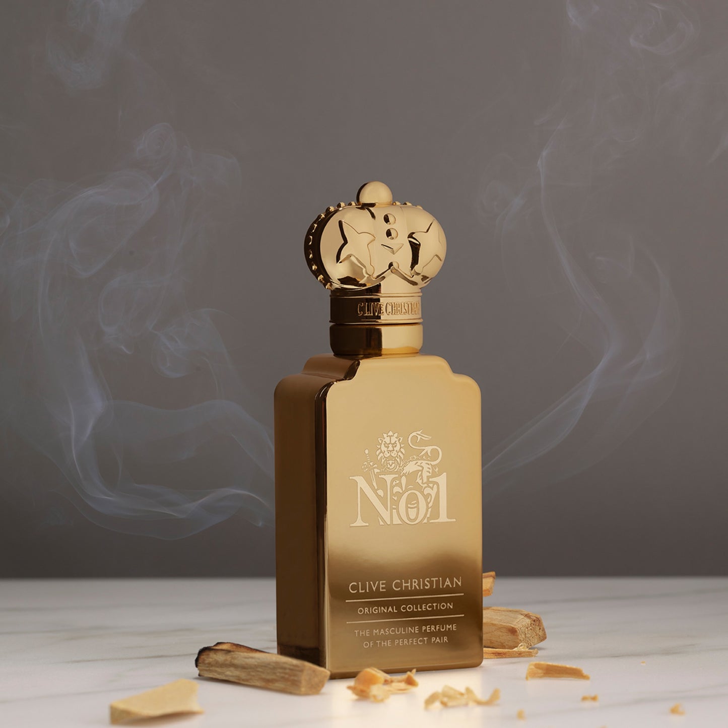 A gold Clive Christian No. 1 perfume bottle stands on a marble surface with sandalwood pieces, wisps of smoke creating an air of luxury.